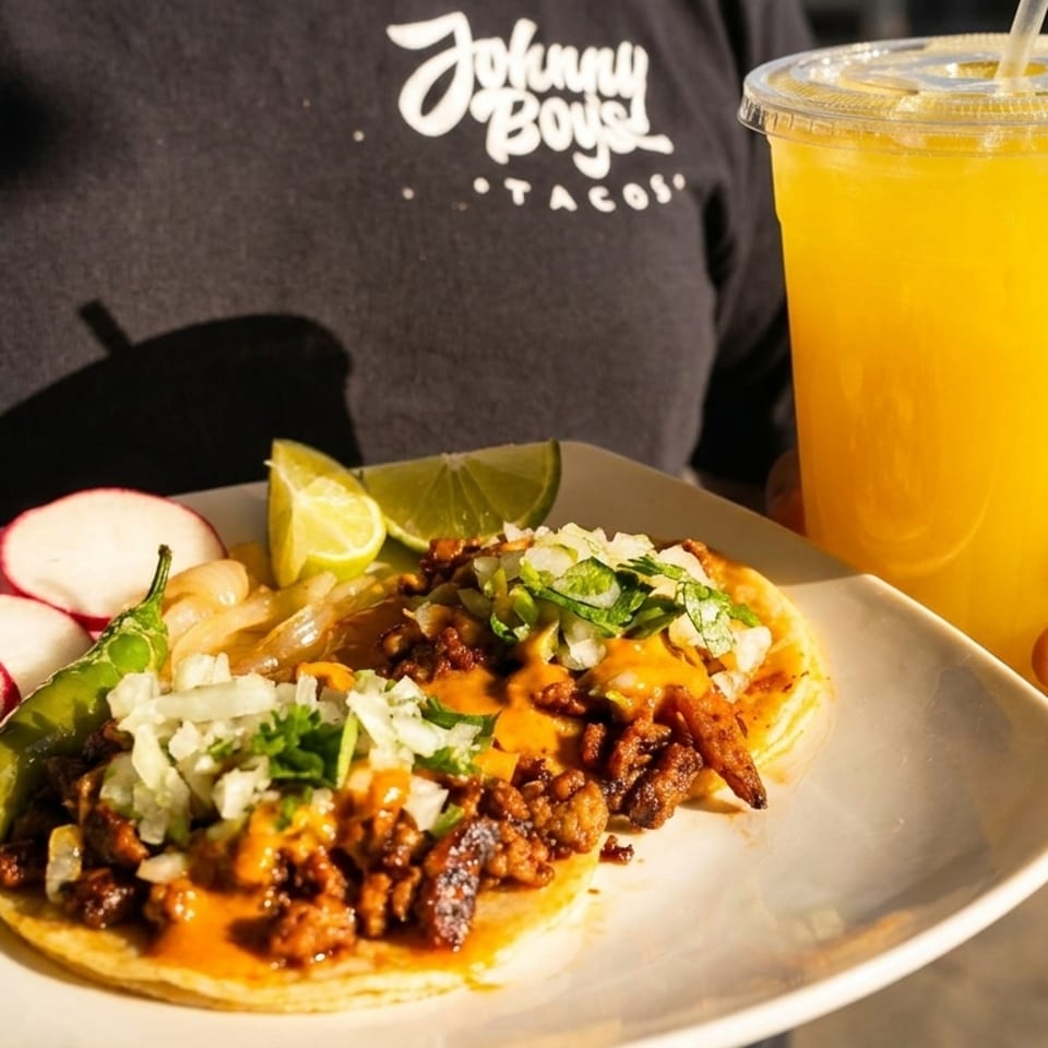 Plate of topped tacos with radishes, grilled pepper, and lime beside a large orange drink, held by a Johnny Boys Tacos staff member.